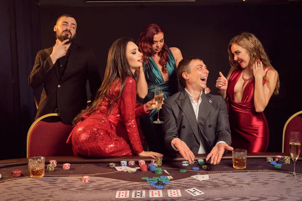 楽観的な友人はカジノでポーカーをしています 彼らは暗い背景の下でテーブルにポーズをとっている間 彼らの勝利を祝い 笑顔で 非常に興奮しています カード チップ アルコール ギャンブル エンターテイメントの概念 — ストック写真