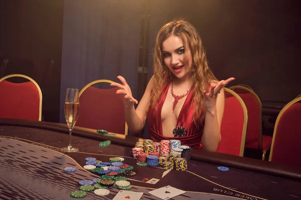 長い巻き毛と完璧な化粧 セクシーな赤いドレスに身を包んだきれいな女性 ギャンブルのテーブルに座り笑顔で勝利を喜ぶ 黄色のスポットライトの線で暗い煙の背景にポーカーの概念 カジノ — ストック写真