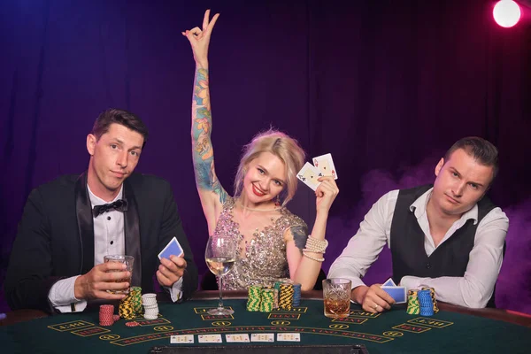 Two stately men and pretty maiden are playing poker at casino. Youth are making bets waiting for a big win. They are smiling and looking at the camera sitting at the table against a red and blue backlights on black smoke background. Cards, chips, mon