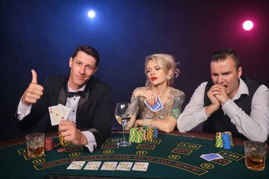 İki zengin adam ve çekici bayan kumarhanede poker oynuyorlar. Gençler büyük bir galibiyet için bahis oynuyorlar. Siyah dumanlı arka planda kırmızı ve mavi arka farlara karşı masada otururken başparmak gösteriyorlar. Kartlar, fişler, para, kumar,