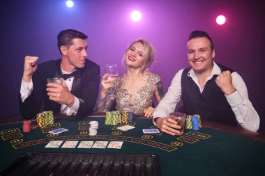 İki yakışıklı erkek ve harika bir kız kumarhanede poker oynuyorlar. Gençler büyük bir galibiyet için bahis oynuyorlar. Masada oturmuş gülümsüyor ve poz veriyorlar. Siyah duman arka planında kırmızı ve mavi bir arka ışığa karşı. Kartlar, çipler, para, kumar