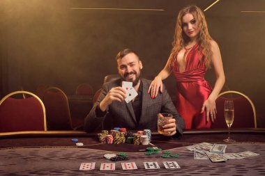 Yakışıklı erkek ve çekici kadın kumarhanede poker oynuyorlar. Gençler kazanma kombinasyonunu sergiliyorlar ve masada sigara dumanına karşı poz verirken seviniyorlar. Kartlar, çipler, para, kumar, eğlence konsepti.