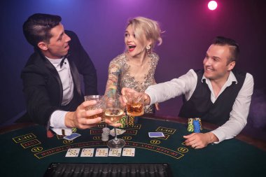 İki yakışıklı erkek ve seksi kadın kumarhanede poker oynuyorlar. Gençler büyük bir galibiyet için bahis oynuyorlar. Masada oturmuş gülümsüyor ve poz veriyorlar. Siyah duman arka planında kırmızı ve mavi bir arka ışığa karşı. Kartlar, fişler, para, kumar, ent