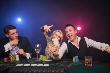 İki atletik erkek ve çekici bayan kumarhanede poker oynuyorlar. Gençler büyük bir galibiyet için bahis oynuyorlar. Masada oturmuş gülümsüyor ve poz veriyorlar. Siyah duman arka planında kırmızı ve mavi bir arka ışığa karşı. Kartlar, çipler, para, kumar