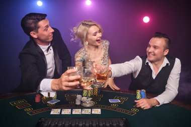İki yakışıklı erkek ve seksi kadın kumarhanede poker oynuyorlar. Gençler büyük bir galibiyet için bahis oynuyorlar. Siyah duman arka planında kırmızı ve mavi arka farlara karşı masada oturmuş, gülümsüyor ve alkol içiyorlar. Kartlar, çipler, para, 