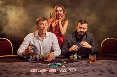 İki yakışıklı arkadaş ve seksi kız kumarhanede poker oynuyorlar. Büyük bir galibiyet için bahis oynuyorlar, gülümsüyorlar ve masada arka planda sarı bir ışığa karşı poz verirken iyi vakit geçiriyorlar. Kartlar, fişler, para, kumar, en