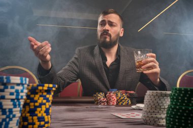 Klasik gri takım elbiseli görkemli sakallı erkek kumarhanede poker oynuyor. Masada fişler ve kartlarla oturuyor. Elinde bir bardak viski tutuyor ve birilerine bakıyor. Kumar bağımlılığı. Samimi duygular ve eğlence.