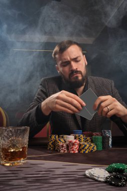 Esmer sakallı, klasik gri ceketli bir erkek kumarhanede poker oynuyor. Üzerinde cips ve bir bardak viskiyle masada oturuyor. O, elindeki kartlara bakıyor. Kumar bağımlılığı. Eğlence konsepti.