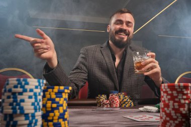 Klasik gri takım elbiseli zeki sakallı adam kumarhanede poker oynuyor. Üzerinde fişler ve kartlarla masada oturuyor. Elinde bir bardak viski tutuyor ve birini işaret ediyor. Kumar bağımlılığı. Samimi duygular ve eğlence adamları.