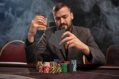 Klasik gri ceketli Brunet sakallı adam kumarhanede duman içinde poker oynuyor üzerinde cipslerle masada oturuyor. Elinde bir bardak viski tutarken elindeki kartlara bakıyor. Kumar bağımlılığı. Eğlence konsepti.