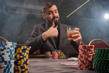 Klasik gri takım elbiseli atletik sakallı adam kumarhanede poker oynuyor. Üzerinde fişler ve kartlarla masada oturuyor. Elinde bir bardak viski tutuyor ve kameraya bakarken başparmağını kaldırıyor. Kumar bağımlılığı. Evet.