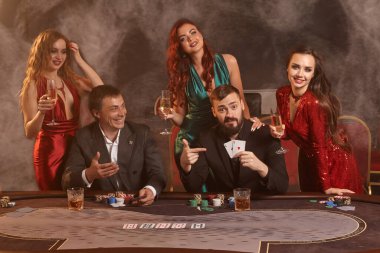 Keyifli ortaklar kumarhanede poker oynuyor. Galibiyetlerini kutluyorlar, gülümsüyorlar ve masada karanlık bir duman geçmişine karşı poz verirken çok heyecanlı görünüyorlar. Kartlar, çipler, para, alkol, kumar, eğlence konsepti.