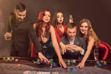 Komik ortaklar kumarhanede poker oynuyorlar. Galibiyetlerini kutluyorlar, gülümsüyorlar ve masada karanlık bir duman geçmişine karşı poz verirken çok heyecanlı görünüyorlar. Kartlar, çipler, para, alkol, kumar, eğlence konsepti.