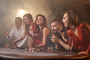 Heyecanlı arkadaşlar kumarhanede poker oynuyorlar. Gençler büyük bir galibiyet için iddiaya girerken masada siyah bir dumanla beyaz bir spot ışığına karşı poz veriyorlar. Kartlar, çipler, para, alkol, kumar, eğlence konsepti.