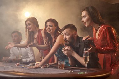 Neşeli meslektaşlar kumarhanede poker oynuyorlar. Gençler büyük bir galibiyet için iddiaya girerken masada siyah bir dumanla beyaz bir spot ışığına karşı poz veriyorlar. Kartlar, çipler, para, alkol, kumar, eğlence konsepti.
