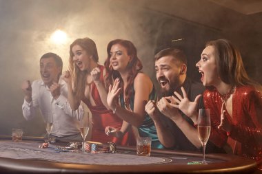 Heyecanlı sınıf arkadaşları kumarhanede poker oynuyorlar. Gençler büyük bir galibiyet için iddiaya girerken masada siyah bir dumanla beyaz bir spot ışığına karşı poz veriyorlar. Kartlar, çipler, para, alkol, kumar, eğlence konsepti.