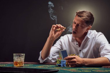 Beyaz gömlekli atletik adam kumarhanedeki masada beyaz bir spot ışığına karşı duman içinde poker oynuyor. O, elindeki kartlara bakıyor ve bir puro içiyor. Kumar bağımlılığı. Samimi duygular ve eğlence konsepti.