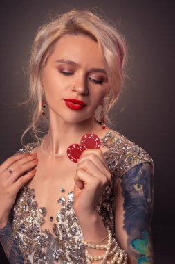 Sarışın, dövmeli ve mükemmel makyajlı, gümüş rengi parlak bir elbise giymiş elinde kırmızı kumar fişleri ile poz veren çekici bir kızın yakın plan fotoğrafı. Siyah arka planda poker konsepti arka ışığa karşı. Kumarhane.