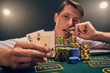 Beyaz gömlekli iyi görünümlü birinin yakın plan çekimi, kumarhanedeki masada duman içinde, beyaz bir spot ışığına karşı poker oynamaktır. Zaferine sevinerek elinde iki as ve ağzında bir çip tutuyor. Kumar bağımlılığı. Evet.