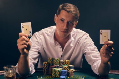 Beyaz gömlekli yakışıklı adam kumarhanenin masasında beyaz bir spot ışığına karşı duman içinde poker oynuyor. Elindeki iki ası göstererek zaferine seviniyor. Kumar bağımlılığı. Samimi duygular ve eğlence konsepti.