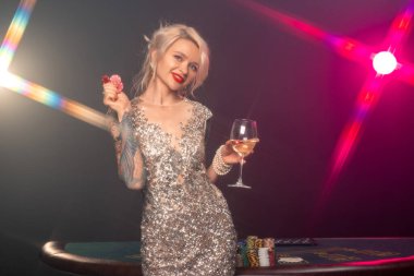 Sarışın, dövmeli, mükemmel makyajlı, gümüş rengi parlak bir elbise giymiş çekici bir kadının orta boy yakın çekimi. Elinde kırmızı cipsler ve elinde bir bardak şampanyayla kumar masasının karşısında duruyor ve gülümsüyor. Poker konsepti