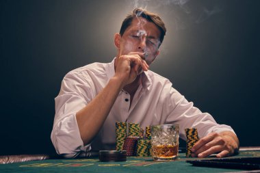 Beyaz gömlekli şık bir adam kumarhanenin masasında beyaz bir spot ışığına karşı duman içinde poker oynuyor. O bir puro içerken ve ona bakarken zaferine seviniyor. Kumar bağımlılığı. Samimi duygular ve eğlence konsepti.