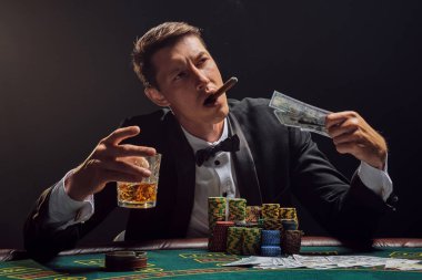 Siyah ve beyaz tişörtlü yakışıklı bir erkek kumarhanedeki masada beyaz bir spot ışığına karşı duman içinde poker oynuyor. Puro içerken, alkol içerken ve elinde biraz para tutarken zaferine seviniyor. Kumar oynuyorum.