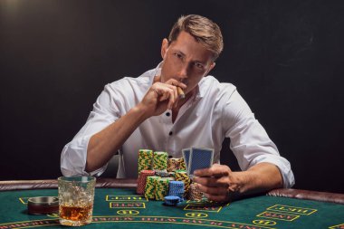 Beyaz gömlekli şık bir adam kumarhanedeki masada beyaz bir spot ışığına karşı duman içinde poker oynuyor. O bir puro içiyor ve elinde bazı kartlar tutuyor. Kumar bağımlılığı. Samimi duygular ve eğlence konsepti.