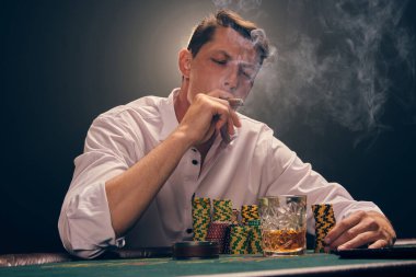 Beyaz gömlekli şık bir adam kumarhanedeki masada beyaz bir spot ışığına karşı duman içinde poker oynuyor. O bir puro içerken ve ona bakarken zaferine seviniyor. Kumar bağımlılığı. Samimi duygular ve eğlence konsepti.