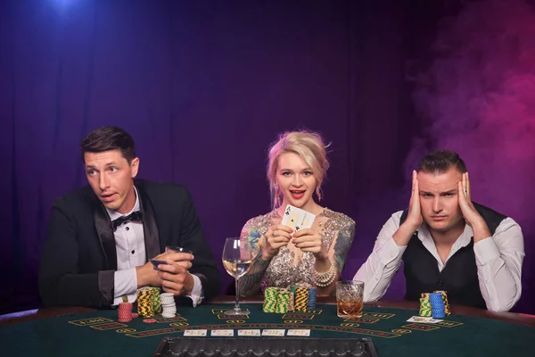 2人の威厳のある男と可愛い女の子がカジノでポーカーをしています 若者は大きな勝利を待って賭けをしています 黒い煙の背景に赤と青のバックライトに向かってテーブルに座って笑顔とポーズをとっています カード チップ ギャンブル — ストック写真