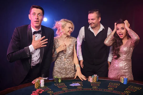 4人の興奮した友人のグループがカジノでポーカーをしています 若者は大きな勝利を待って賭けをしています 彼らは黒の背景に赤と青のバックライトに対してテーブルの上に立っている カード チップ ギャンブル エンターテイメントの概念 — ストック写真