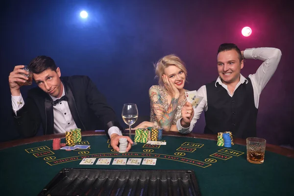 Two elegant men and attractive maiden are playing poker at casino. Youth are making bets waiting for a big win. They are smiling and looking at the camera while sitting at the table against a red and blue backlights on black smoke background. Cards,