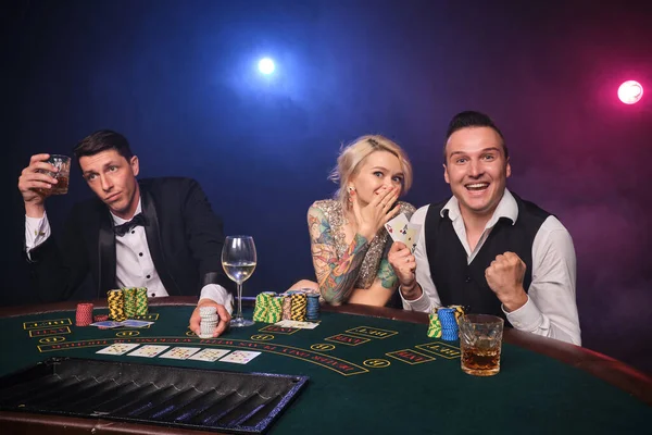 2人のエレガントな仲間と魅力的な女性がカジノでポーカーをしています 若者は大きな勝利を待って賭けをしています 黒い煙の背景に赤と青のバックライトに向かってテーブルに座って笑顔とポーズをとっています カード チップ ギャンブル — ストック写真