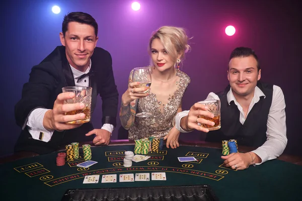 Two good-looking men and amazing woman are playing poker at casino. Youth are making bets waiting for a big win. They are smiling and drinking champagne sitting at the table against a red and blue backlights on black smoke background. Cards, chips, m