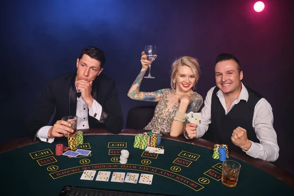 Two elegant men and attractive woman are playing poker at casino. Youth are making bets waiting for a big win. They are smiling and drinking alcohol while sitting at the table against a red and blue backlights on black smoke background. Cards, chips,