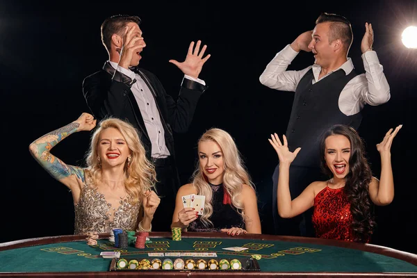 幸せな友人はカジノでポーカーをしています 彼らは 白いスポットライトの光の中で暗い背景を背景にテーブルにポーズを取りながら 彼らの勝利 笑顔を祝い 非常に興奮しています カード チップ アルコール ギャンブル — ストック写真