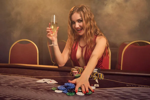 長い巻き毛と完璧なメイクアップを持つ女性を魅了し セクシーな赤いドレスを着ています 彼女はギャンブルのテーブルに座っていて チップとシャンパンを持っています 黄色のスポットライトの線で暗い煙の背景にポーカーの概念 カジノ — ストック写真