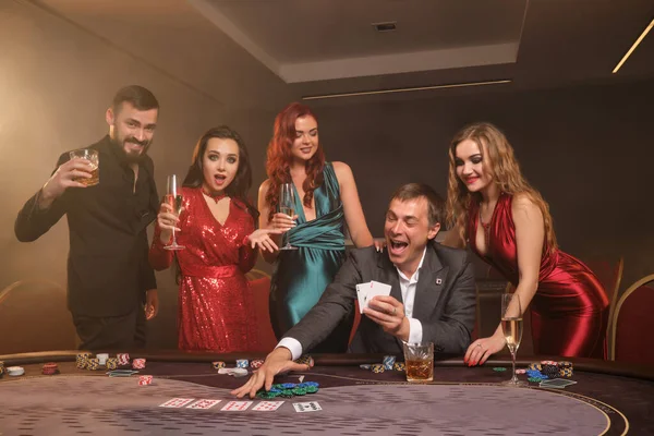 熱狂的なクラスメートはカジノでポーカーをしています 彼らは 暗闇の煙の背景にスポットライトを当てながらテーブルにポーズをとりながら 彼らの勝利を祝い 笑顔で 非常に興奮しています カード チップ アルコール ギャンブル — ストック写真