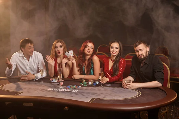 裕福なパートナーはカジノでポーカーをしています 彼らは 暗闇の煙の背景にスポットライトを当てながらテーブルにポーズをとりながら 彼らの勝利を祝い 笑顔で 非常に興奮しています カード チップ アルコール ギャンブル — ストック写真