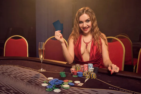 美丽的女人 一头长长的卷发 完美的妆容 穿着性感的红色连衣裙 她坐在赌桌旁 手里拿着两张扑克牌 面带微笑 扑克的概念在黑暗的烟幕背景在一束黄色的聚光灯下 Casi — 图库照片