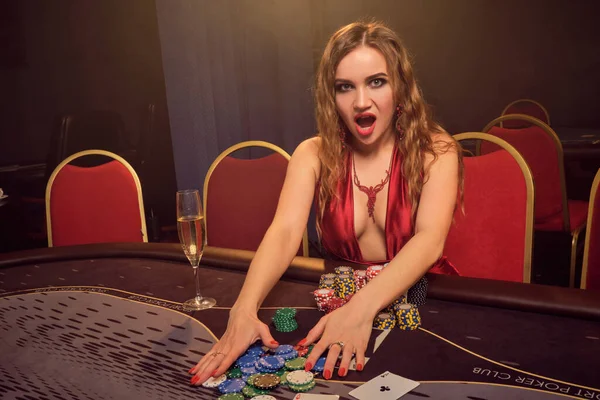 長い巻き毛と完璧な化粧 セクシーな赤いドレスに身を包んだきれいな女の子 ギャンブルのテーブルに座って笑っている間に 彼女は勝利を喜び 疑問に思っています 黄色のスポットライトの線で暗い煙の背景にポーカーの概念 — ストック写真