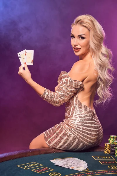 Close-up shot of a beautiful girl with blond hair and bright make-up, dressed in a golden striped shiny dress, posing sideways with playing cards in her hands while sitting on a table with chips on it and smiling. Poker concept on a black smoke backg