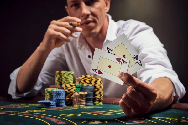 Athletic man in a white shirt is playing poker sitting at the table at casino in smoke, against a white spotlight. He is smoking a cigar while showing two aces in his hand. Gambling addiction. Sincere emotions and entertainment concept.
