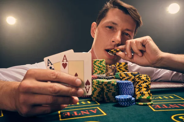 Beyaz gömlekli iyi görünümlü birinin yakın plan çekimi, kumarhanedeki masada duman içinde, beyaz bir spot ışığına karşı poker oynamaktır. Zaferine sevinerek elinde iki as ve ağzında bir çip tutuyor. Kumar bağımlılığı. Evet.