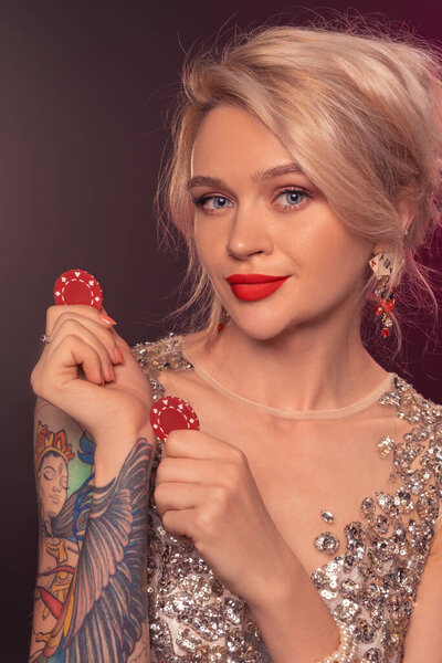 Крупный план очаровательной леди с светлыми волосами, татуированными руками и идеальным макияжем, одетой в серебряное блестящее платье, позирующей с красными фишками в руках и улыбающейся. Концепция покера на черном фоне с подсветкой. Казино
.