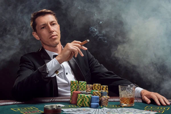 Smart person in a black slassic suit and white shirt is playing poker sitting at the table at casino in smoke, against a white spotlight. He rejoicing his victory smoking a cigar and looking at the camera. Gambling addiction. Sincere emotions and ent