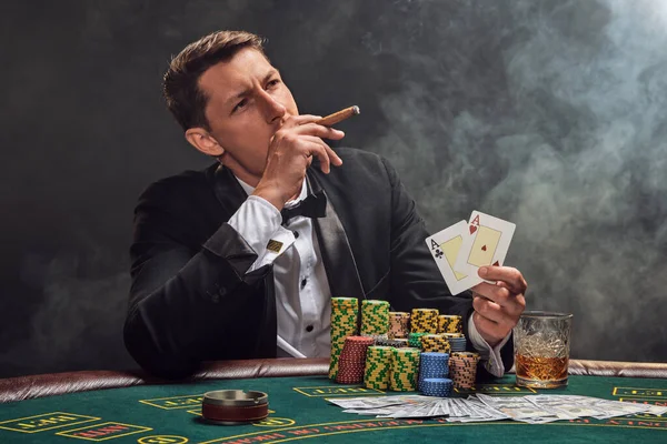 Elegant fellow in a black slassic suit and white shirt is playing poker sitting at the table at casino in smoke, against a white spotlight. He rejoicing his victory showing two aces in his hand and smoking a cigar. Gambling addiction. Sincere emotion