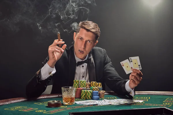 Siyah ve beyaz gömlekli yakışıklı bir erkek kumarhanedeki masada beyaz bir spot ışığına karşı duman içinde poker oynuyor. Elindeki iki ası göstererek, puro içerek ve kameraya bakarak zaferine seviniyor. Kumar. 
