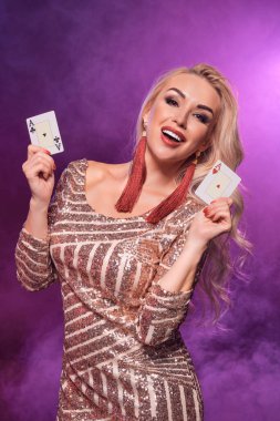 Mükemmel saç stili ve parlak makyajlı, altın rengi parlak bir elbise giymiş, göz kamaştırıcı sarışın bir kızın yakın plan fotoğrafı elinde kartlarla kameraya bakıyor. Siyah duman arka planında pembe ve mavi b ile poker konsepti
