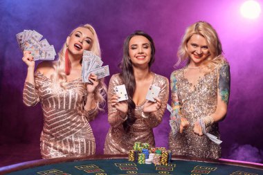 Charming women with a perfect hairstyles and bright make-up, dressed in a golden shiny dresses are posing standing at a gambling table and looking happy. Poker concept on a black smoke background with pink and blue backlights. Casino. clipart